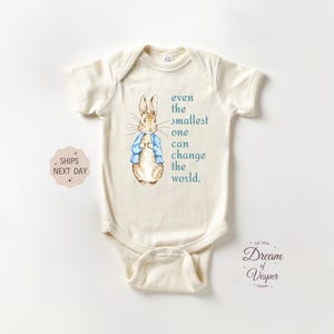 May include: Cream-colored baby onesie featuring a watercolor illustration of a rabbit wearing a blue jacket. The text "even the smallest one can change the world" is printed on the front. The onesie has short sleeves and a snap closure.