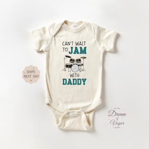 May include: Cream-colored baby bodysuit with the text "Can't wait to jam with Daddy" and a drum set graphic. The text and drum set are in teal and black. The bodysuit is made of soft material.