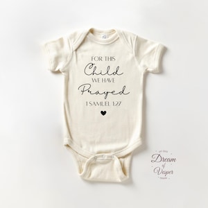May include: Cream-colored baby onesie with the text "FOR THIS Child WE HAVE Prayed 1 SAMUEL 1:27" in black script. A small black heart is below the text. The onesie has short sleeves and a snap closure.