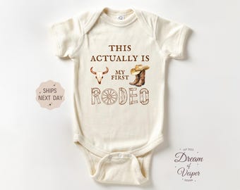 May include: Cream-colored baby bodysuit with the text "THIS ACTUALLY IS MY FIRST RODEO" in brown. The design includes a steer skull, cowboy boots, and a wagon wheel. A cute outfit for a baby.