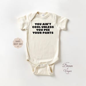 May include: Cream-colored baby onesie with the text "YOU AIN'T COOL UNLESS YOU PEE YOUR PANTS" in black. The onesie has short sleeves and a snap closure. A small logo and text are visible in the lower right corner.