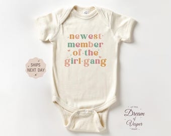 May include: Cream-colored baby bodysuit with the text "newest member of the girl gang" in colorful lettering. The bodysuit has short sleeves and a snap closure. The design includes small hearts.