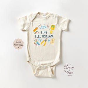 May include: Cream-colored baby onesie with the text "Tiny Electrician" and illustrations of tools like a screwdriver, pliers, and a lightbulb. The design includes blue lightning bolts and a plug.