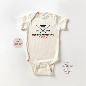 May include: Cream-colored baby onesie with a black and red graphic design. The design includes a Japanese torii gate, crossed swords, and the text "EST. 2025 HONOR'S APPRENTICE JASON" in black and red. The onesie is short-sleeved.