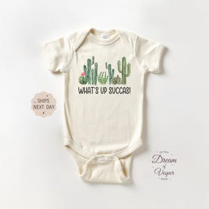May include: Cream-colored baby bodysuit with a cactus graphic and the text "WHAT'S UP SUCCAS!". The design features various green cacti and a pink flower. The bodysuit is made of soft material.