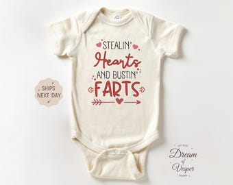 May include: Cream-colored baby onesie with the text "STEALIN' Hearts AND BUSTIN' FARTS" in red. The design includes hearts and arrows. A small logo is in the bottom right corner.
