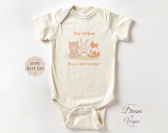May include: Cream-colored baby bodysuit with the text "The Littlest Book Club Member" and a graphic of a bear, bunny, and fox. The design is in soft orange tones. The bodysuit is short-sleeved.