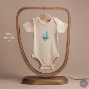 May include: Cream-colored baby onesie with the name "Jason" printed in black script with a teal "J". The onesie is hanging on a wooden hanger within a wooden display stand. The stand has a curved design and a wooden base.