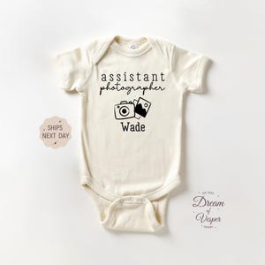 May include: Cream-colored baby bodysuit with the text "assistant photographer" above a camera and photo icon, and the name "Wade" below. The bodysuit is short-sleeved and has a snap closure.