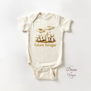 May include: Cream-colored baby bodysuit with a mushroom illustration and the text "future forager" in gold. The design features a cluster of detailed mushrooms. A charming and nature-inspired baby garment.