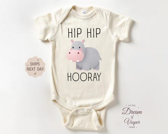 May include: Cream-colored baby bodysuit with the text "HIP HIP HOORAY" and a cartoon hippo illustration. The hippo is gray with a pink snout. The bodysuit has short sleeves and a snap closure.