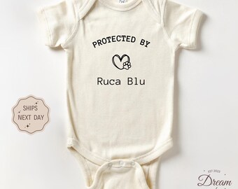May include: Cream-colored baby onesie with the text "PROTECTED BY" above a heart and paw print design, followed by "Ruca Blu" in black lettering. The onesie is a short-sleeved garment, perfect for newborns and infants.