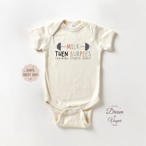 May include: Cream-colored baby onesie with the text "MILK THEN BURPEES TRAINING STARTS EARLY" in a playful font. The design includes a barbell graphic. A small logo is visible in the lower right corner.