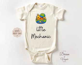 May include: Cream-colored baby bodysuit with a colorful cartoon cat mechanic graphic. The cat wears a blue hat and holds tools. Text reads "little Mechanic". A small design and text "Dream of Vesper Design" are also visible.