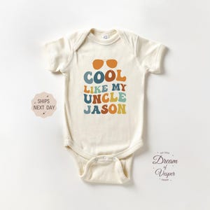 May include: Cream-colored baby bodysuit with the text "COOL LIKE MY UNCLE JASON" in retro-style lettering. The design includes orange sunglasses above the text. A small logo and text are in the lower right corner.