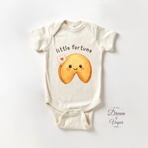May include: Cream-colored baby onesie with a cartoon fortune cookie graphic. The fortune cookie has a smiling face and a small heart-shaped note. The text "little fortune" is above the cookie.
