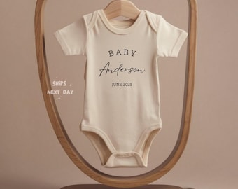May include: Cream-colored baby onesie with the text "BABY Anderson JUNE 2025" in black script. The onesie hangs on a wooden hanger within a decorative wooden frame. The overall aesthetic is minimalist and modern, perfect for a baby announcement.