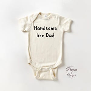 May include: Cream-colored baby bodysuit with the text "Handsome like Dad" in black. The short-sleeved onesie has a round neckline and snap closure at the bottom. The Dream of Vesper Design logo is in the bottom right corner.
