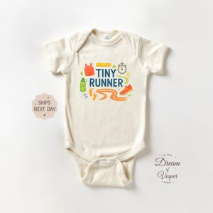 May include: Cream-colored baby onesie with a colorful "Tiny Runner" graphic. The design includes a stopwatch, running shoes, a water bottle, and a running track. The onesie is made of soft material.
