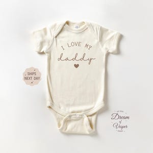 Peut inclure: Body bébé crème à manches courtes. Le texte "I LOVE MY daddy" est imprimé en marron, avec un petit cœur sous le mot "daddy". Le body est fait d'une matière douce.