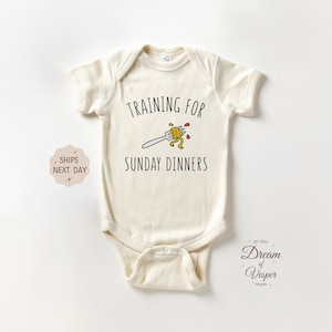 May include: Cream-colored baby onesie with the text "TRAINING FOR SUNDAY DINNERS" above a fork with spaghetti. The onesie has short sleeves and a snap closure. The design includes a small illustration of a fork with spaghetti.