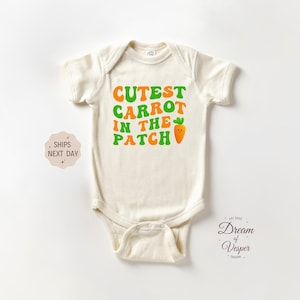 May include: A cream-colored baby onesie with the text "CUTEST CARROT IN THE PATCH" in green and orange letters. A cartoon carrot with a smiley face is next to the text.
