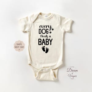 May include: Cream-colored baby bodysuit with the text "every DOG Needs a BABY" in black. The design includes a paw print and baby footprints. The bodysuit has short sleeves and a snap closure.