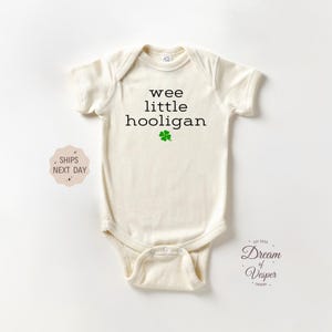 May include: Cream-colored baby onesie with the text "wee little hooligan" in black, and a green shamrock. The onesie has short sleeves and a snap closure. The design is centered on the front of the garment.