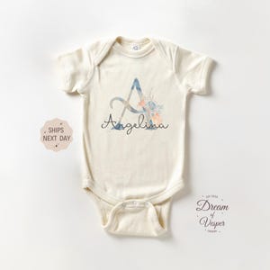 May include: Cream-colored baby onesie with a watercolor floral design. The letter "A" is featured with the name "Angelina" in script. The onesie has short sleeves and a snap closure.