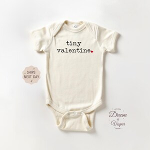 May include: Cream-colored baby bodysuit with the text "tiny valentine" in black, a small red heart, and short sleeves. The bodysuit is made of soft material and has a snap closure at the bottom. The design is simple and suitable for Valentine's Day.