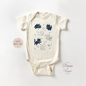 May include: Cream-colored baby bodysuit featuring a navy blue illustration of cats in space suits, planets, and stars. The text "Dream of Vesper Design" is printed on the lower right. The bodysuit has short sleeves and a snap closure.