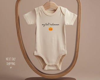 May include: Cream-colored baby bodysuit with short sleeves, featuring the text "my first halloween" above a small orange pumpkin graphic. The bodysuit is hanging on a wooden hanger.