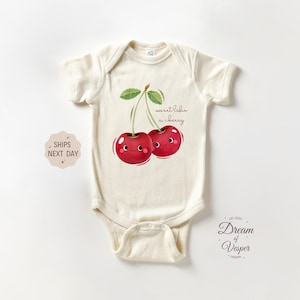 May include: Cream-colored baby bodysuit with a cherry graphic. The illustration features two smiling red cherries with green leaves and the text "sweet like a cherry". The bodysuit is made of soft material.