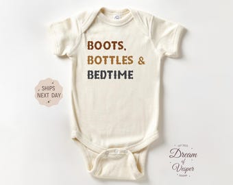 May include: Cream-colored baby bodysuit with the text "BOOTS, BOTTLES & BEDTIME" in brown and navy blue. The short-sleeved onesie has a snap closure at the bottom and a round neckline. The design includes a small logo and text.