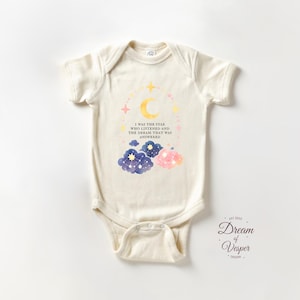 May include: Cream-colored baby onesie with a celestial design. The design includes a crescent moon, stars, and watercolor clouds in blue and pink. Text reads "I WAS THE STAR WHO LISTENED AND THE DREAM THAT WAS ANSWERED."