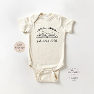 May include: Cream-colored baby bodysuit with short sleeves. The front features the text "second edition" above an open book illustration, with "published 2026" below. A small design and text are visible on the lower right.