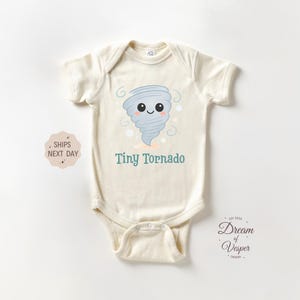 May include: Cream-colored baby bodysuit with a cartoon tornado graphic. The illustration features a smiling gray tornado with the words "Tiny Tornado" in teal. The bodysuit is short-sleeved and has a snap closure.