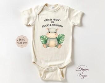 May include: Cream-colored baby onesie with a hippo graphic and the text "HUNGRY HUNGRY FOR SNACKS & SNUGGLES!". The hippo is gray with green leaves. The onesie has short sleeves and a snap closure.
