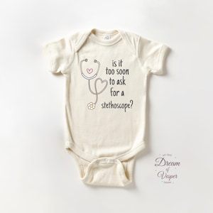 May include: Cream-colored baby onesie with a stethoscope graphic and the text "is it too soon to ask for a stethoscope?" The design includes a pink heart and a small flower. The onesie is a newborn or infant garment.