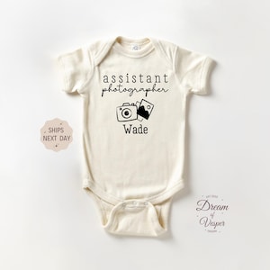 May include: White baby bodysuit with black text that reads "assistant photographer" and a cartoon camera with film. The name "Wade" is printed below the camera.