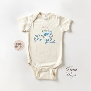 May include: Cream-colored baby onesie with a whimsical airplane graphic. The design features a bunny pilot in a blue plane, with the words "first time flayer" and the name "Anderson" in blue script. A cute and unique baby garment.