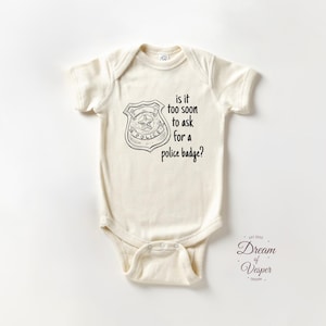 May include: Cream-colored baby onesie with a police badge graphic and the text "is it too soon to ask for a police badge?" The onesie has short sleeves and a snap closure at the bottom.
