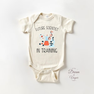 May include: Cream-colored baby onesie with the text "FUTURE SCIENTIST IN TRAINING" and a graphic of lab equipment. The graphic includes a beaker, microscope, and DNA strand. The onesie is short-sleeved.