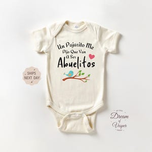May include: A cream-colored baby onesie with a black graphic that reads "Un Pajarito Me Dijo Que Van a Ser Abuelitos" with a pink heart and a blue bird on a branch.