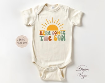 May include: Cream baby bodysuit with the text "HERE COMES THE SON" in a retro font, featuring a yellow sun graphic. The design includes orange, green, and pink lettering. A cute and stylish outfit for a baby.