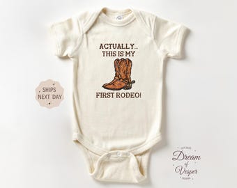 May include: Cream-colored baby onesie with a brown cowboy boot graphic and the text "ACTUALLY... THIS IS MY FIRST RODEO!". The onesie has short sleeves and a snap closure.
