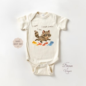 May include: Cream-colored baby bodysuit with a cartoon raccoon illustration. The raccoon is running, with paint spills in yellow, orange, and blue. Text reads "I came, I saw, I made a mess."