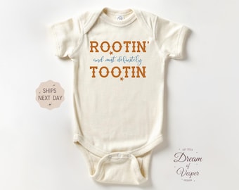 May include: Cream-colored baby onesie with the text "ROOTIN' and must definitely TOOTIN" in brown and blue. The onesie has short sleeves and a snap closure. The design includes star accents.