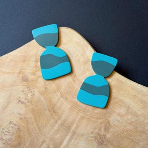 May include: Two pairs of turquoise and gray abstract earrings. Each earring has a top section and a bottom section, with a gray stripe in the middle. The earrings are displayed on a wooden surface.