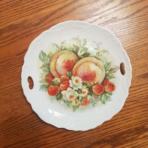 May include: White ceramic serving plate with a scalloped edge and two handles. The plate features a painted design of peaches, strawberries, and white flowers.